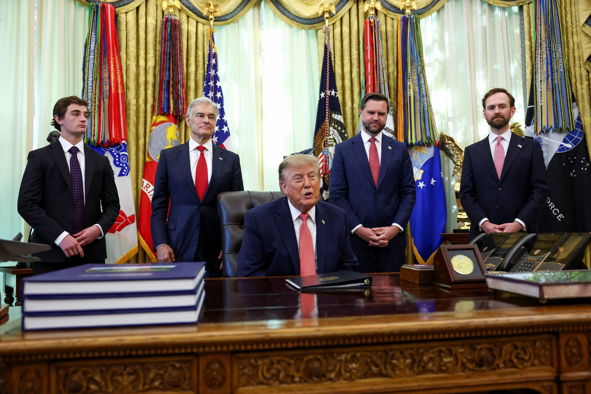 President Donald Trump seated at the Resolute Desk in the Oval Office, with four men in suits, including Vice President JD Vance, standing behind him in front of military flags.